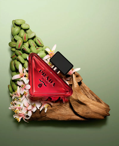 Red Prada triangle-shaped perfume bottle with green beans and flowers on a green background
